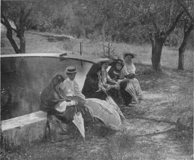 There are six ladies in this photo, arranged informally seated on one side of a short stone wall that appears to surround a small pool of water, with several trees in the background. Three of the ladies are dressed in light colored Western attire that completely covers their bodies, with light-colored brimmed hats. The other three ladies are in dark robes with attached head coverings. These three are holding folded umbrellas with their hands. The three in dark attire appear to be of late teen or early adult age, with the other three appearing to be somewhat older.
